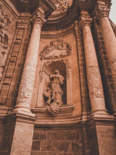 A detailed stone sculpture depicting a robed figure standing in a niche, surrounded by ornate carvings and flanked by grand columns. The rich textures of the stone and intricate details of the artwork are highlighted, creating a sense of historical and architectural grandeur.