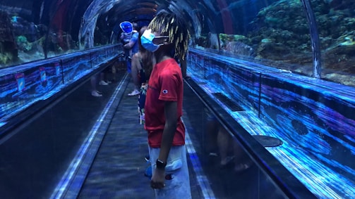 Inside an underwater tunnel at an aquarium, a young person wearing a mask stands on a moving walkway, surrounded by large tanks showcasing marine life. Reflections of blue lights and water patterns create an immersive experience as other visitors explore the tunnel.