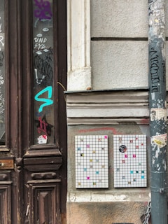 An urban street scene with two mosaic-style art pieces mounted on a worn wall. The artwork consists of white grid panels with scattered colorful squares and designs. Surrounding the art, the area is filled with graffiti on old wooden doors and a pole. The atmosphere is gritty and vibrant, with a mix of textures from the weathered surfaces and lively street art.