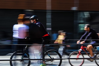 Cyclists and pedestrians in motion create a dynamic and blurred scene on a city street, conveying speed and movement. People are captured with a sense of urgency as they navigate the urban environment.