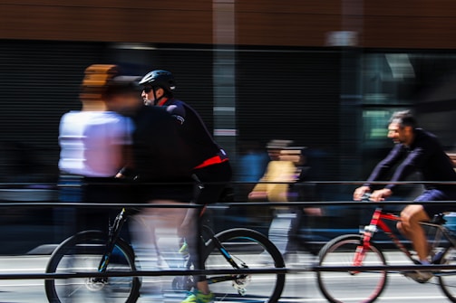 Cyclists and pedestrians in motion create a dynamic and blurred scene on a city street, conveying speed and movement. People are captured with a sense of urgency as they navigate the urban environment.