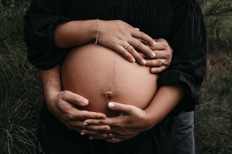 A close-up of a pregnant belly being gently held by two sets of hands. One hand set, adorned with a bracelet and a ring, rests above, while another set of larger hands with a wedding band cradles from below. The background features soft greenery, creating a serene and natural atmosphere.