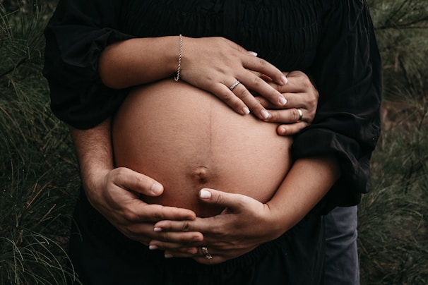 A close-up of a pregnant belly being gently held by two sets of hands. One hand set, adorned with a bracelet and a ring, rests above, while another set of larger hands with a wedding band cradles from below. The background features soft greenery, creating a serene and natural atmosphere.