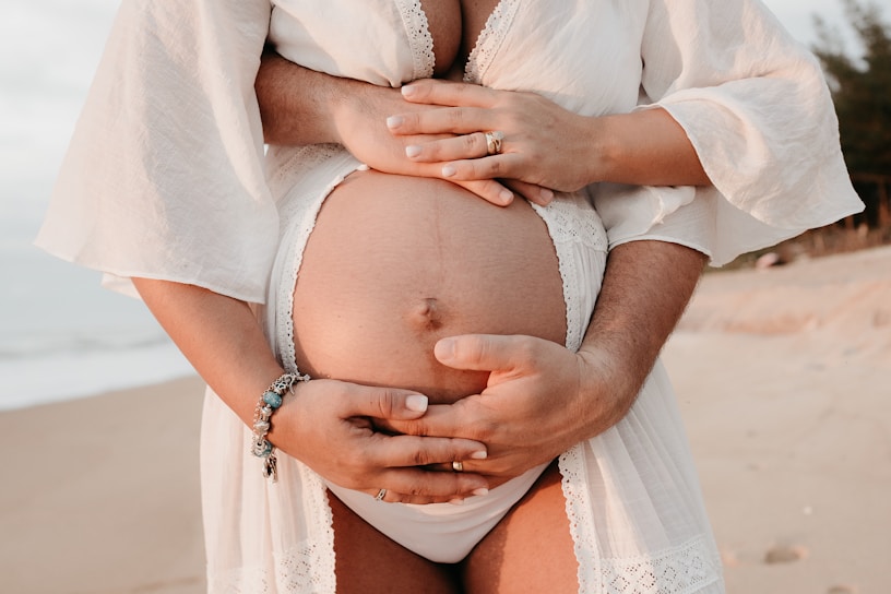 A pregnant belly is embraced by two pairs of hands, one from the person carrying the baby and the other from a supporting individual behind. The setting appears to be a beach, as sand is visible in the background. The person is wearing a white flowing garment, and there is a bracelet on one wrist.