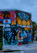 A vibrant street mural featuring a variety of colorful elements, including a person with sunglasses and a red dress lounging, a silhouette of a person on a bicycle, fruit patterns, palm trees, and architectural elements. The mural is located on the corner of a building and includes text reading 'Wynwood Walls 305'.
