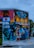 A vibrant street mural featuring a variety of colorful elements, including a person with sunglasses and a red dress lounging, a silhouette of a person on a bicycle, fruit patterns, palm trees, and architectural elements. The mural is located on the corner of a building and includes text reading 'Wynwood Walls 305'.