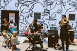 A group of musicians performing outside against a wall decorated with abstract line art. The band consists of a keyboard player, a trombone player, a conga player, and two other percussionists. The setting suggests a street performance, with musical equipment set up on the ground.
