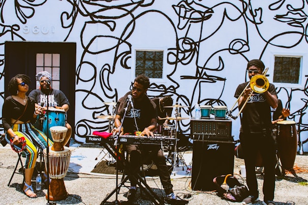 A group of musicians performing outside against a wall decorated with abstract line art. The band consists of a keyboard player, a trombone player, a conga player, and two other percussionists. The setting suggests a street performance, with musical equipment set up on the ground.