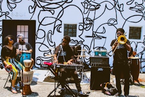 A group of musicians performing outside against a wall decorated with abstract line art. The band consists of a keyboard player, a trombone player, a conga player, and two other percussionists. The setting suggests a street performance, with musical equipment set up on the ground.
