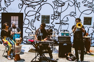 A group of musicians performing outside against a wall decorated with abstract line art. The band consists of a keyboard player, a trombone player, a conga player, and two other percussionists. The setting suggests a street performance, with musical equipment set up on the ground.