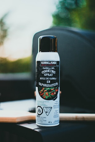 A can of Kirkland brand canola oil cooking spray placed on a dark surface. The can has a white body with a black cap, and the label features a photo of vegetables being cooked in a pan. The background is slightly blurred with hints of green and light colors, suggesting an outdoor setting with natural lighting.