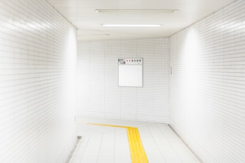 A white-tiled hallway with a simple, clean design. The walls and ceiling are covered in small, square tiles, and there's a bright yellow tactile guidance path on the floor leading into the distance. A sign with text hangs on one of the walls.