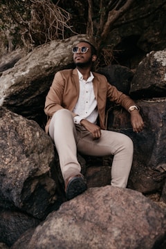 A person is sitting on large rocks wearing a brown blazer, white shirt, and light-colored pants. They are wearing sunglasses and looking upwards, appearing relaxed and contemplative.