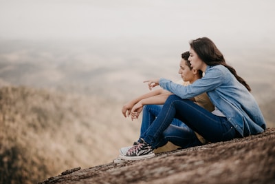 A young couple sits together on a rocky hill, with the woman pointing towards the distance. They appear relaxed and engaged in conversation, with a blurry, expansive landscape in the background.