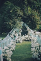 An outdoor wedding aisle adorned with white flowers and greenery leads to an elegant backdrop of draped fabric and floral arrangements. The setting is lush with green grass and surrounded by tall trees.