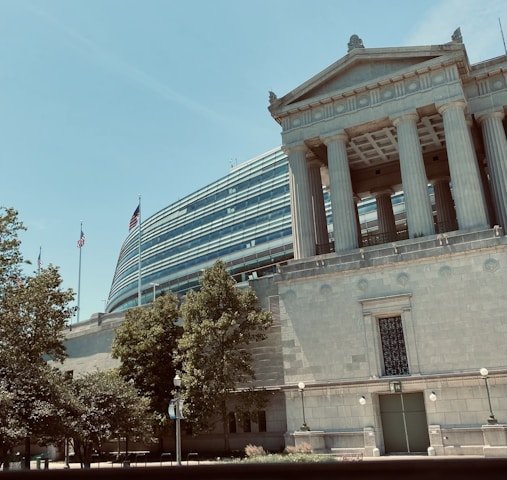 A neoclassical building with large columns stands beside a modern structure with curved, glass-paneling. Several flags are visible, and trees are in the foreground under a clear blue sky.