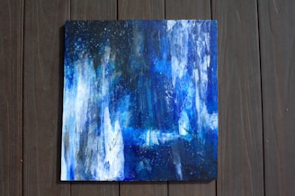 An abstract painting with a mix of deep blue, white, and black colors. The composition consists of textured strokes and splashes, evoking a sense of celestial or underwater scenes. The artwork is placed on a wooden surface, highlighting the contrast between the natural and the abstract.