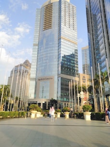 Tall modern skyscrapers with reflective glass facades dominate the scene. The central building bears the name 'The Ritz-Carlton' and is surrounded by lush greenery and a plaza. People are scattered around the area, some standing and others walking. The sky is bright with a few clouds.