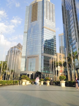 Tall modern skyscrapers with reflective glass facades dominate the scene. The central building bears the name 'The Ritz-Carlton' and is surrounded by lush greenery and a plaza. People are scattered around the area, some standing and others walking. The sky is bright with a few clouds.