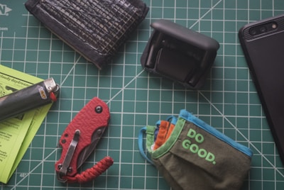 A selection of personal items is arranged on a green gridded cutting mat. There is a black wallet, a black lighter, a red folding knife, a compact drone, and a pouch with 'DO GOOD' printed on it. A portion of a black smartphone is visible on the top right corner.