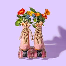 A pair of pink roller skates adorned with vibrant flowers emerging from the tops. The flowers are red, orange, and yellow with green leaves. The wheels of the skates are translucent and pink, set against a light purple background.