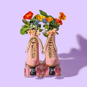 A pair of pink roller skates adorned with vibrant flowers emerging from the tops. The flowers are red, orange, and yellow with green leaves. The wheels of the skates are translucent and pink, set against a light purple background.
