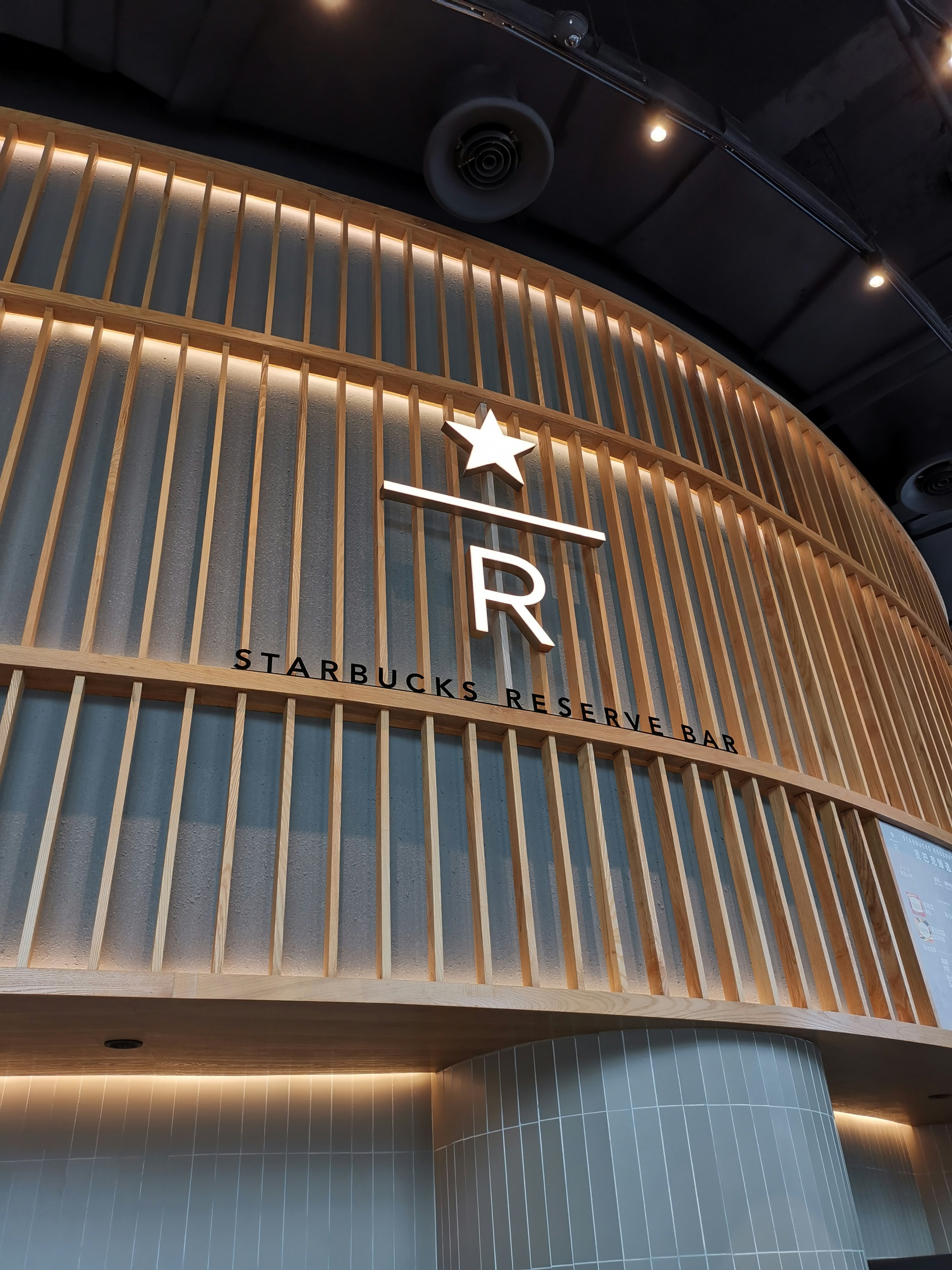 A modern, upscale interior design featuring a prominent wooden slat wall with a large letter 'R' beneath a star. The sign reads 'Starbucks Reserve Bar' under sleek overhead lighting. The architectural style is minimalist with a touch of elegance.