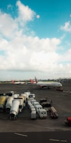 The image depicts an airport tarmac with two large airplanes parked. There are multiple mobile boarding ramps and service vehicles lined up in organized rows. The sky is partly cloudy with a bright, blue color.