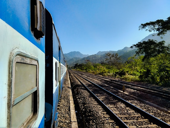 A blue train is traveling along a track surrounded by lush green vegetation, with distant mountains visible under a clear blue sky.