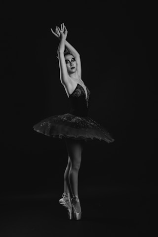A ballerina stands en pointe with her arms gracefully lifted, wearing a dark tutu. The lighting captures her elegant posture and expression, highlighting the contours of the costume and her poised stance.