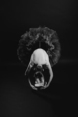 A ballerina is captured in a graceful pose with her arms outstretched towards the ground. She is wearing a detailed, elaborate tutu and ballet shoes, and her head is adorned with a delicate headpiece. The image is in black and white, highlighting the intricate textures and contrasts.