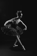 A ballerina is gracefully posing on pointe in a tutu, with a crown on her head. The image is in black and white, creating a dramatic and elegant atmosphere. The lighting highlights her poised form against a dark background.