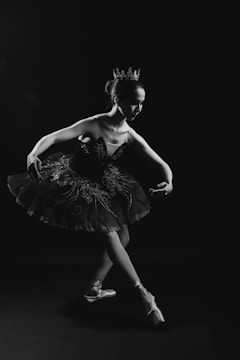 A ballerina is gracefully posing on pointe in a tutu, with a crown on her head. The image is in black and white, creating a dramatic and elegant atmosphere. The lighting highlights her poised form against a dark background.