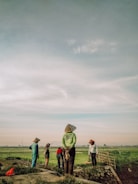 A group of farmers, wearing traditional hats, are standing in a green field under a vast, cloudy sky. They seem to be taking a break or observing the landscape. The morning or evening light casts a warm glow over the scene.