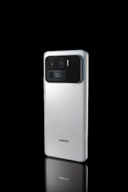 A smartphone with a silvery back and a prominent camera module is positioned against a black background. The phone's rear panel includes dual lenses within the camera setup, and there is text indicating '120X' near the camera. The word 'xiaomi' is clearly visible on the lower part of the phone.