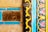 Detailed patterns and textures feature prominently, with intricate floral designs in pink and blue bordered by vibrant yellow and black wavy lines. The rustic elements include a piece of wood with a natural, cracked texture, contrasting with the polished, colorful tiles.