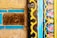 Detailed patterns and textures feature prominently, with intricate floral designs in pink and blue bordered by vibrant yellow and black wavy lines. The rustic elements include a piece of wood with a natural, cracked texture, contrasting with the polished, colorful tiles.