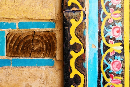 Detailed patterns and textures feature prominently, with intricate floral designs in pink and blue bordered by vibrant yellow and black wavy lines. The rustic elements include a piece of wood with a natural, cracked texture, contrasting with the polished, colorful tiles.