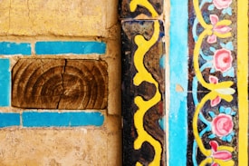 Detailed patterns and textures feature prominently, with intricate floral designs in pink and blue bordered by vibrant yellow and black wavy lines. The rustic elements include a piece of wood with a natural, cracked texture, contrasting with the polished, colorful tiles.