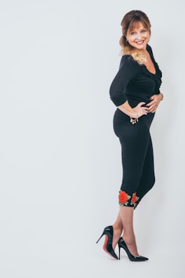 A woman is standing against a plain white background, wearing an all-black outfit with floral embroidery at the bottom of her leggings. She is smiling and looking at the camera, with her hand on her hip. She has long hair styled with loose curls and is wearing black high heels.