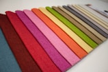 A neatly arranged assortment of fabric swatches displays a broad spectrum of colors, ranging from deep blue and maroon to vibrant pinks and reds, as well as earthy greens, yellows, and browns. The textures are distinct, showcasing a woven pattern on each swatch.