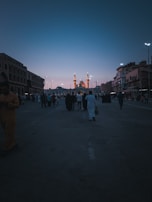 A wide street lined with buildings on both sides converges towards a large, illuminated mosque with multiple minarets. The sky is a gradient from a deep blue to lighter hues near the horizon, suggesting early evening or dawn. Several people wearing traditional clothing walk towards the mosque.