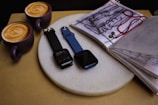 Two smartwatches on a round, white marble surface, accompanied by an open sketchbook and two cups of latte art coffee in purple mugs placed on a brown background.