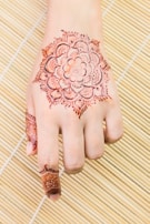 A hand adorned with an intricate henna design, featuring floral and paisley patterns, set against a bamboo mat background. The artwork extends from the back of the hand to the fingers.