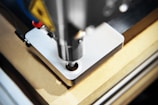 A detailed close-up of a CNC machine that is milling or drilling into a piece of metal on a workbench. The focus is on the cutting tool and the metal piece, showing precision and industrial craftsmanship.