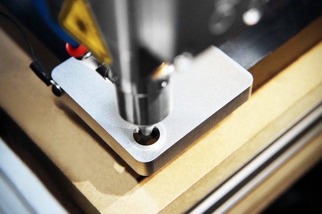 A detailed close-up of a CNC machine that is milling or drilling into a piece of metal on a workbench. The focus is on the cutting tool and the metal piece, showing precision and industrial craftsmanship.