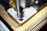 A detailed close-up of a CNC machine that is milling or drilling into a piece of metal on a workbench. The focus is on the cutting tool and the metal piece, showing precision and industrial craftsmanship.