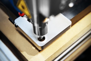 A detailed close-up of a CNC machine that is milling or drilling into a piece of metal on a workbench. The focus is on the cutting tool and the metal piece, showing precision and industrial craftsmanship.