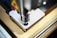A detailed close-up of a CNC machine that is milling or drilling into a piece of metal on a workbench. The focus is on the cutting tool and the metal piece, showing precision and industrial craftsmanship.