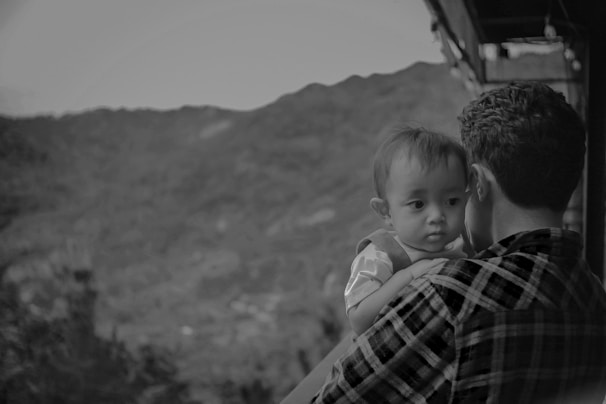 A black and white photograph depicts a man holding a baby. The baby looks outward with a thoughtful expression, as the man, wearing a plaid shirt, gazes at the landscape. They are positioned on a balcony overlooking a mountainous view, creating a serene and intimate scene.